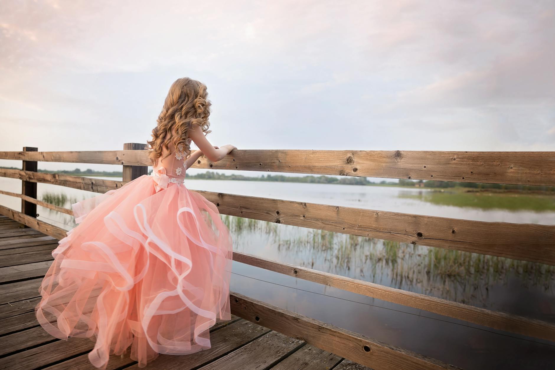 A girl in a flowing pink gown stands on a dock overlooking a serene lake at dusk.