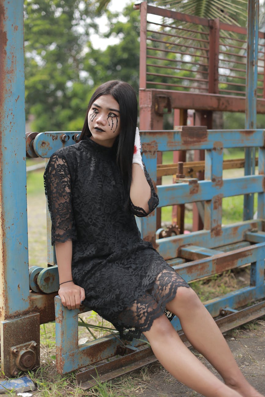 A woman in a black lace dress with creative makeup sits next to a metal railing. [pexels]