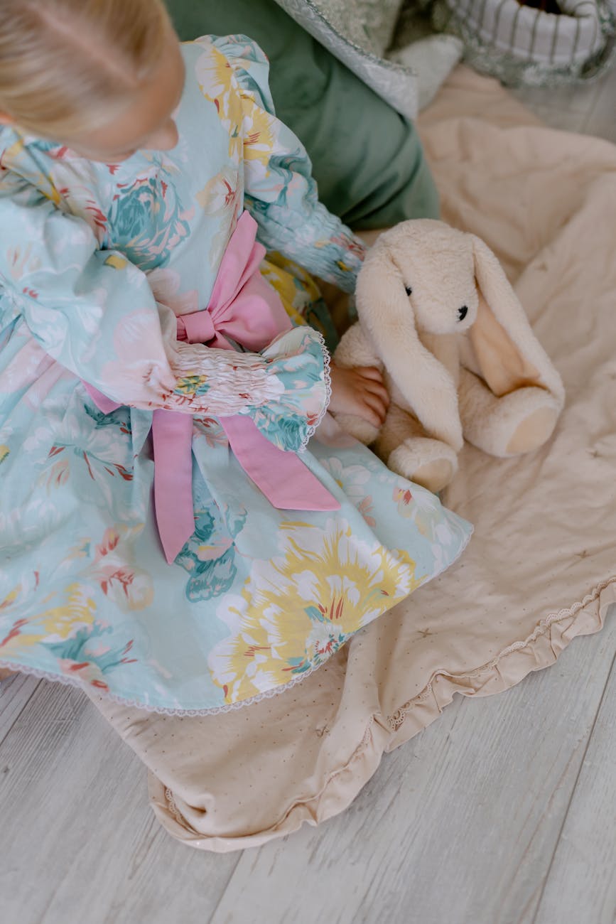 A young girl in a floral dress with a pink bow sits with a plush bunny toy on a cozy beige blanket. [pexels]