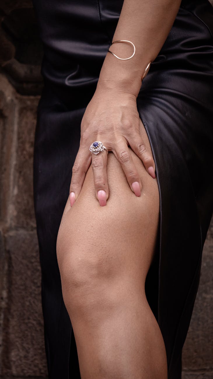 Close-up of a woman's hand with a ring, touching her black leather dress in an elegant setting. [pexels]