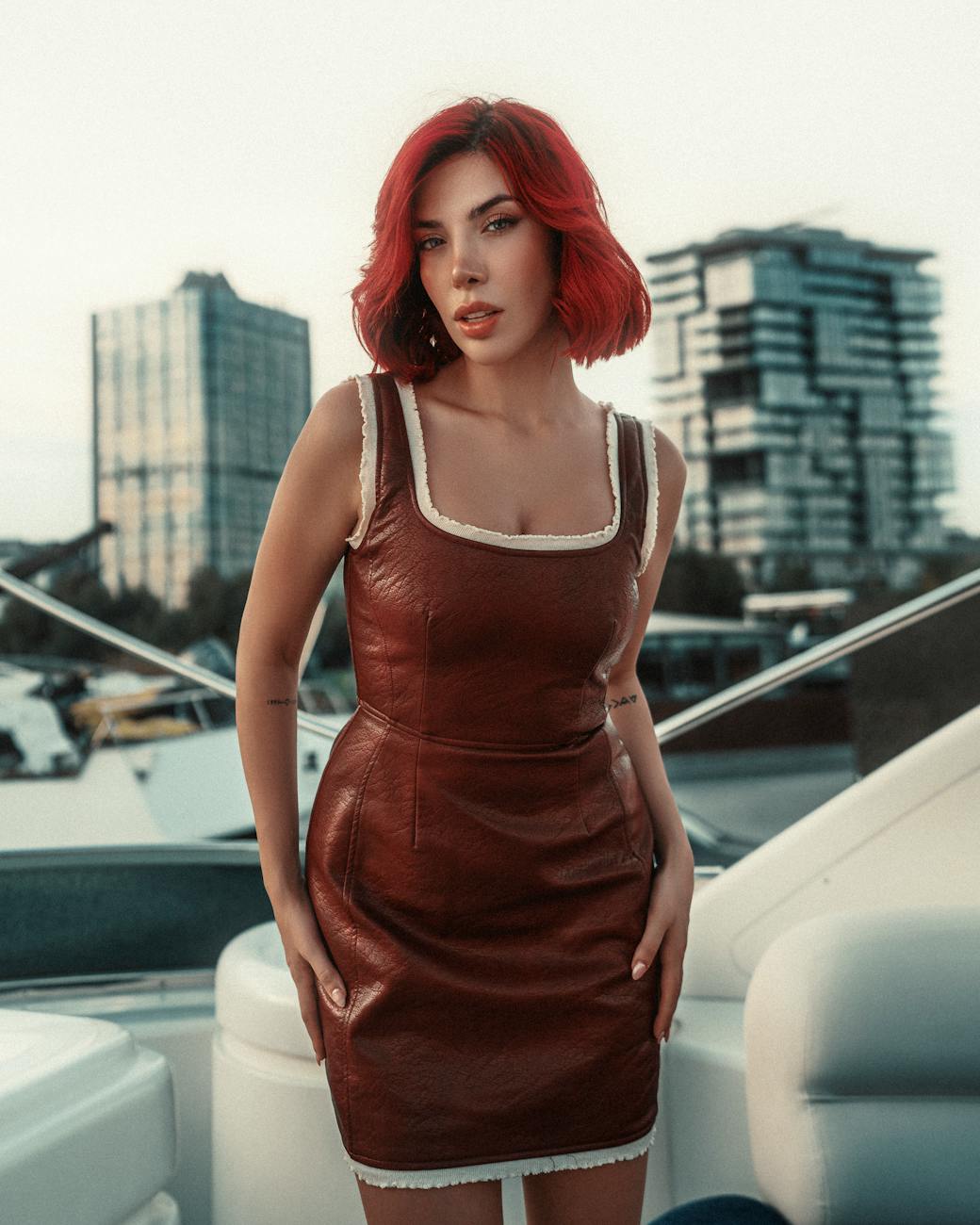 Elegant woman with red hair in leather dress posing confidently on yacht, Dubai skyline in background. [pexels]